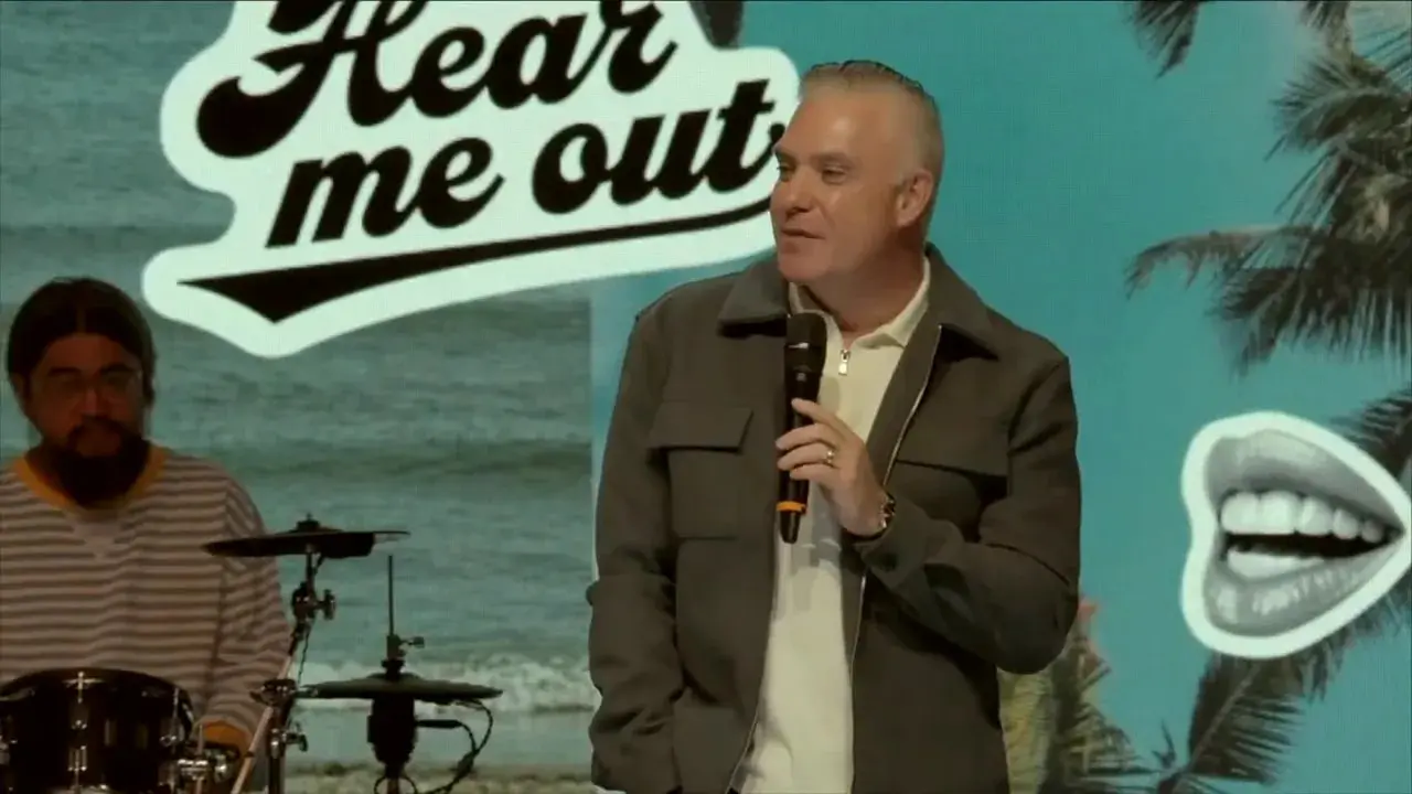 Speaker holding a microphone onstage with a prominent 'Hear me out' logo and palm illustration on the screen behind and a drummer to the left