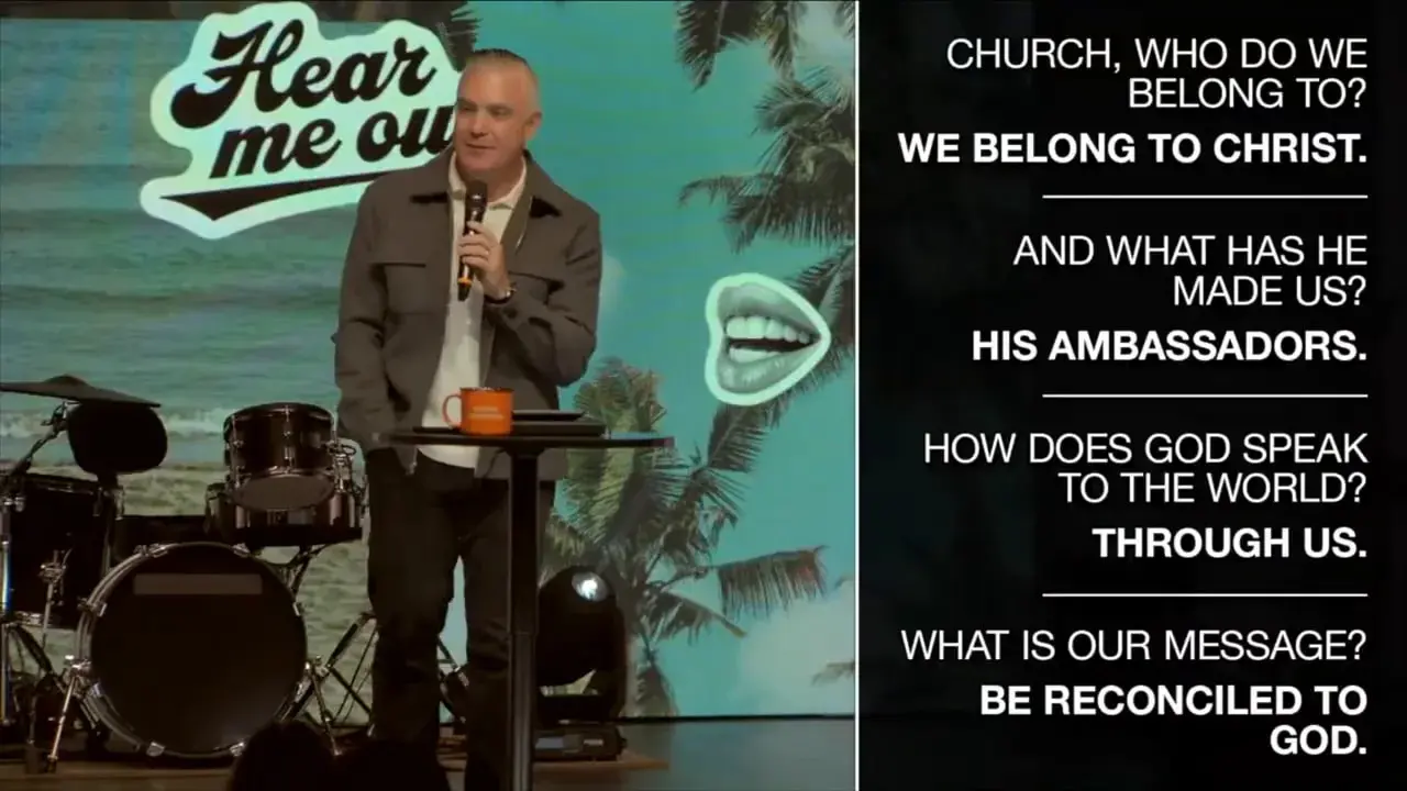 Speaker on stage beside a readable slide stating 'We belong to Christ. His ambassadors. Through us.'