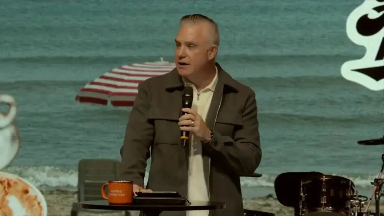 Speaker at a podium holding a microphone with an orange mug and tablet on the table, beach and umbrella backdrop and 'Hear me out' signage