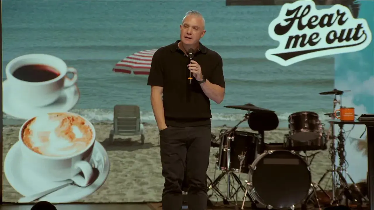 Speaker speaking on stage with 'Hear me out' sign and palm tree backdrop