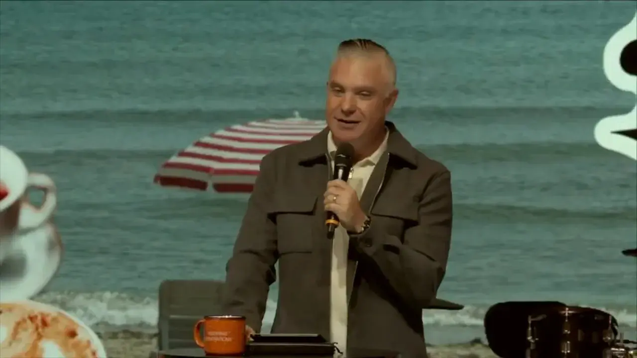 Speaker looking down at notes with one hand on the table near an orange mug, beach backdrop and umbrella